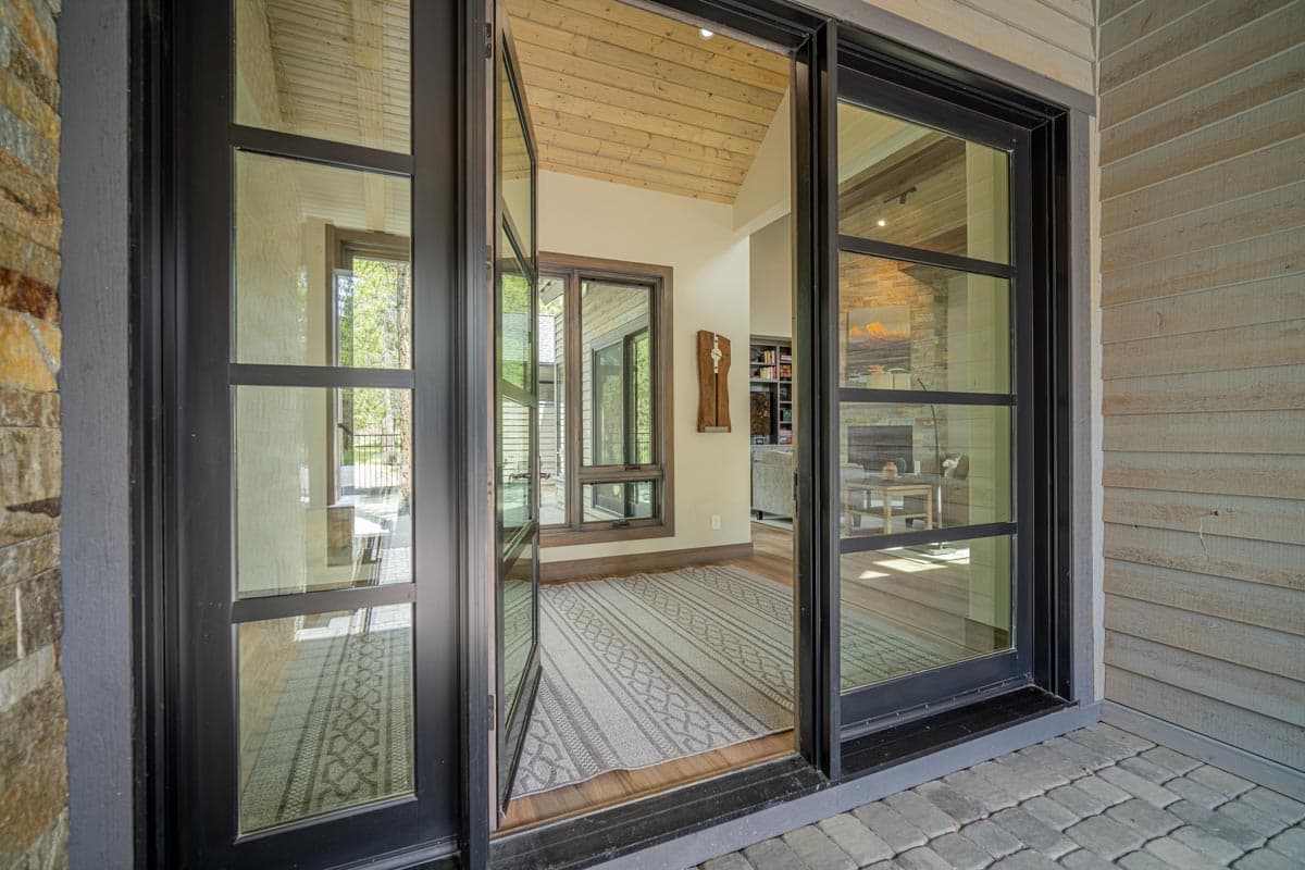 Modern house plan exterior featuring large black-framed glass doors, stone and wood siding, and a paved patio.