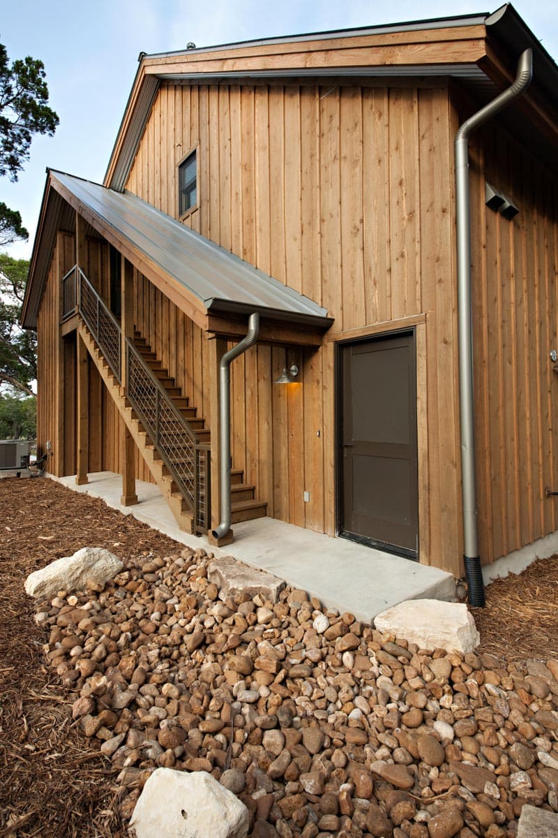 House plan exterior with vertical wood siding, metal roof, exterior stairs, and a dark door.