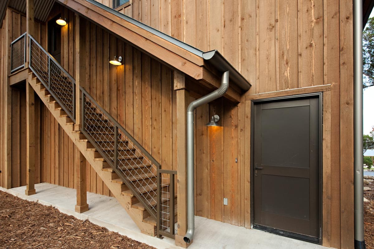 Exterior house plan detail with wooden siding, outdoor stairs with metal mesh railing, and a dark door.