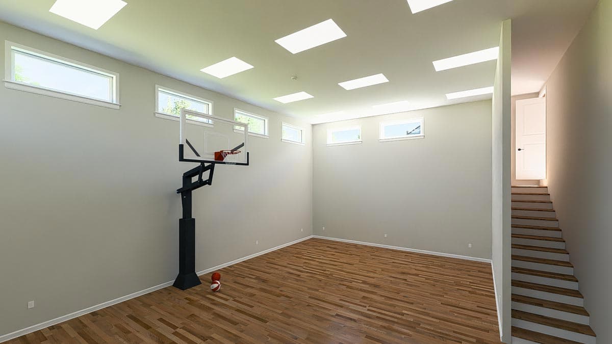 Interior rendering of a recreational room with a basketball hoop, wood flooring, skylights, and a staircase to an open doorway.