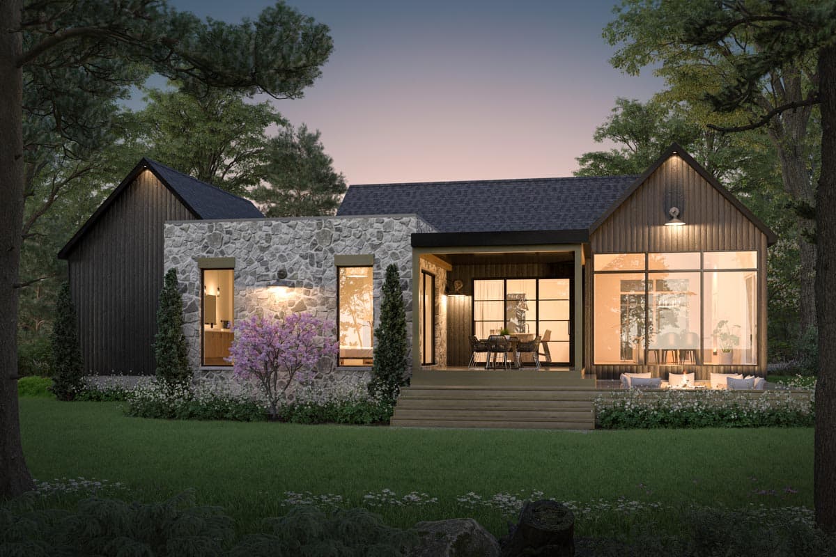 A modern home with stone and wood exterior sits in a wooded area at dusk. Large windows reveal an open interior and an outdoor seating area.