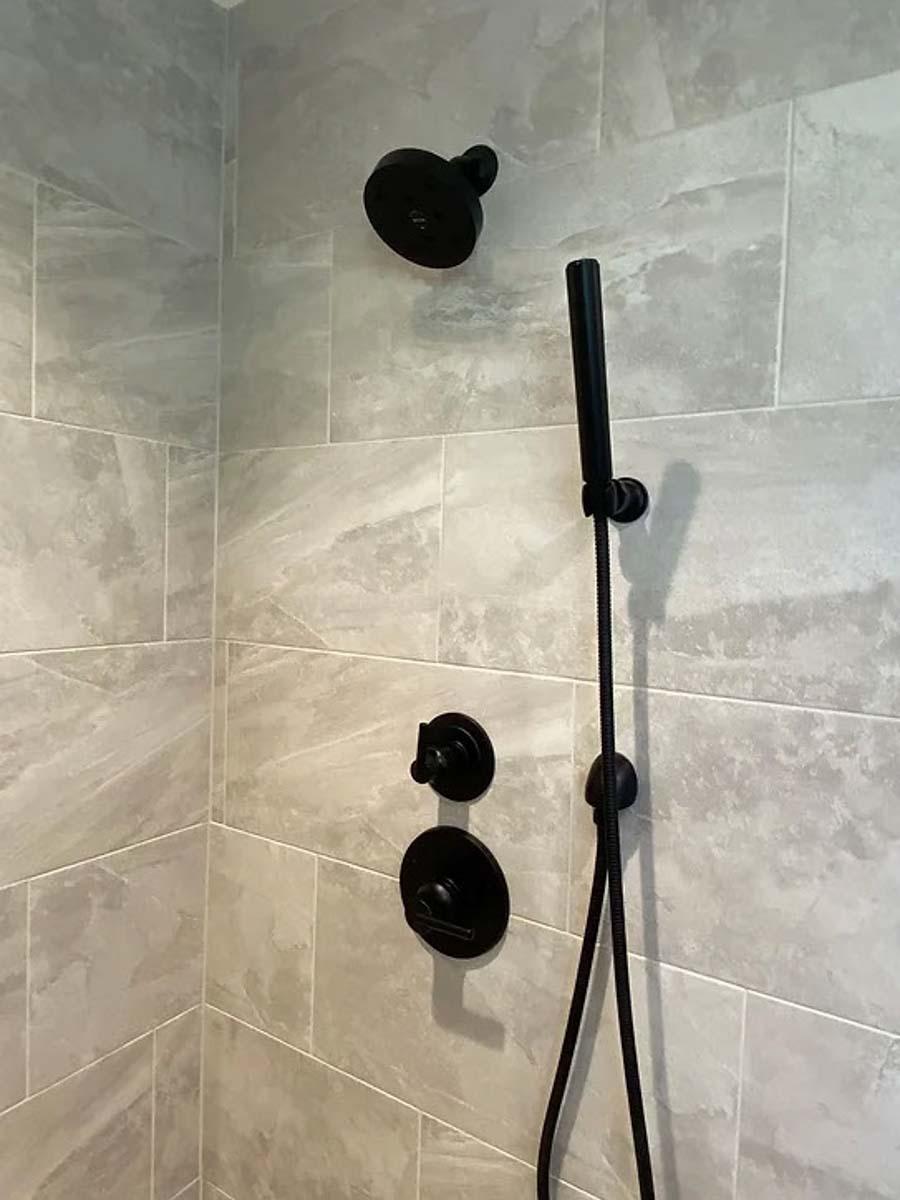 Shower with black fixtures including shower head, hand shower, and controls on gray tiled wall.