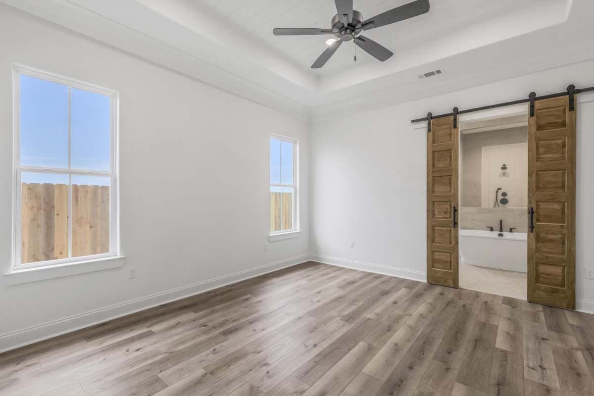 Bedroom with tray ceiling, fan, two windows, and barn doors opening to a bathroom with tub and shower.
