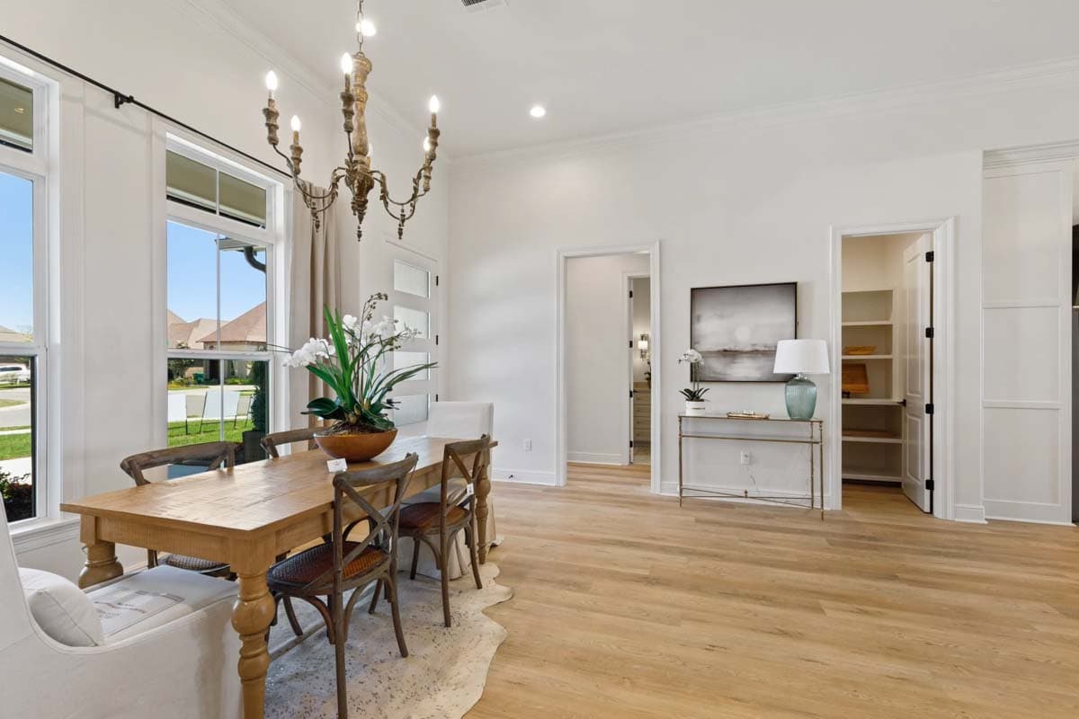 Dining room with wooden table, chairs, ornate chandelier, and large windows overlooking houses.
