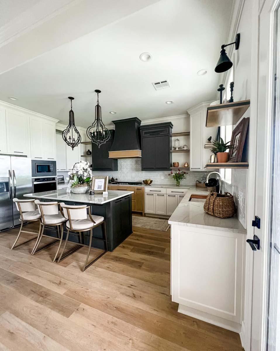 Modern kitchen with a dark island, white cabinetry, stainless steel appliances, and wood plank flooring. An overhead vent hood is featured.