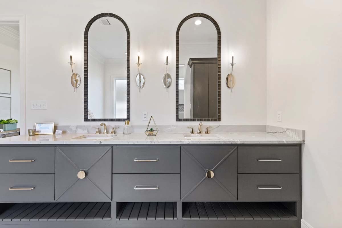 Double vanity with marble countertop, two arched mirrors, brass faucets, and gray cabinetry with open shelving below.