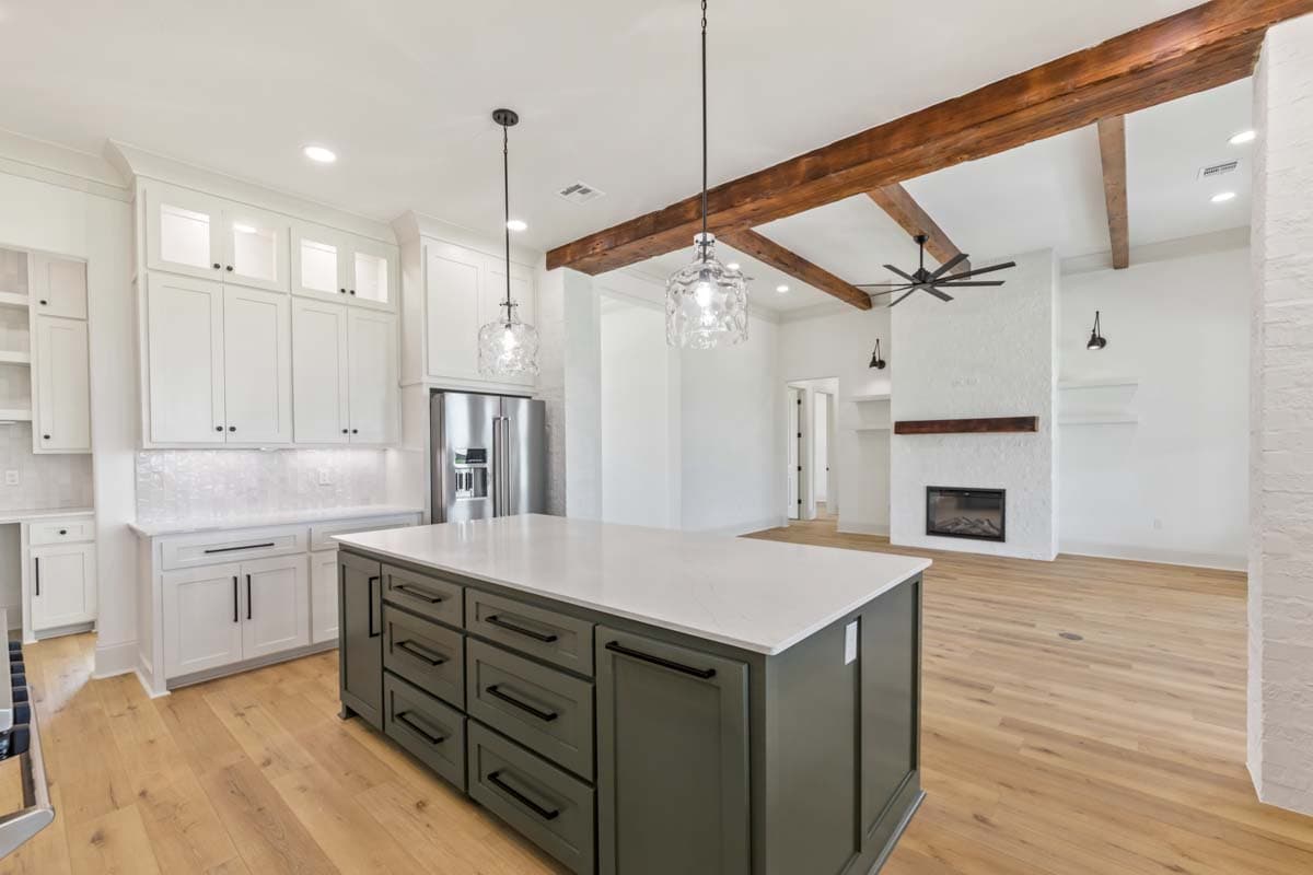 Kitchen with large island, white cabinetry, stainless steel refrigerator, and open view to great room with fireplace and wood beams.