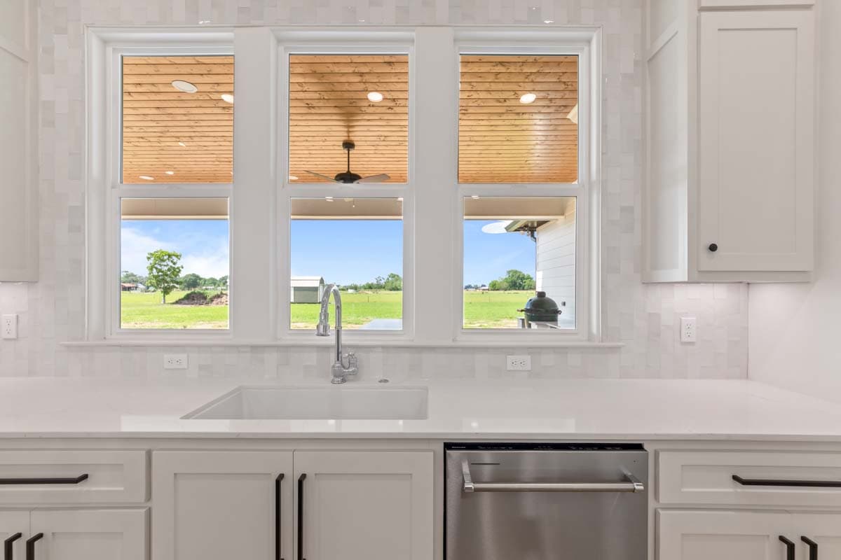Kitchen interior view of sink, dishwasher, and cabinets with a wide window overlooking a covered patio with wood ceiling and outdoor view.