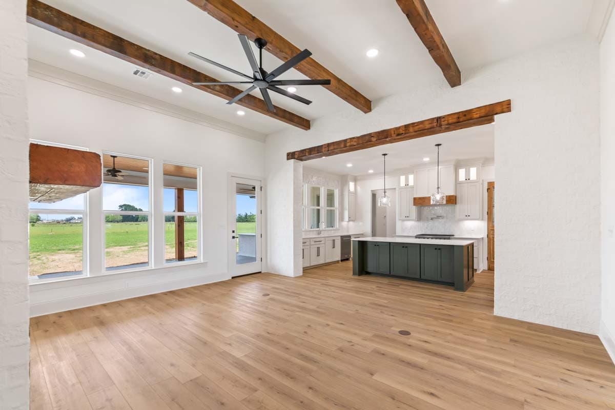 Open-concept living area with exposed wood beams, large windows, and a kitchen with a dark green island and white cabinetry.