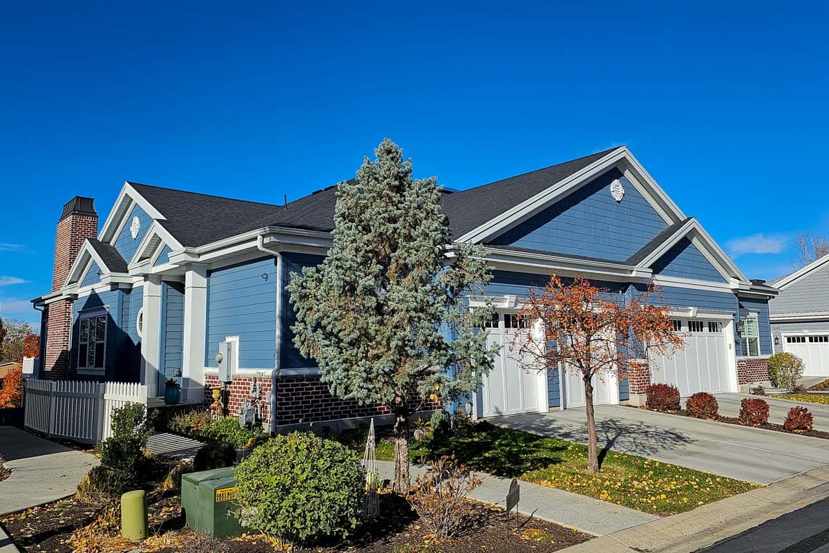 House plan exterior: Blue siding, white trim, brick foundation, prominent gables, and a three-car garage.