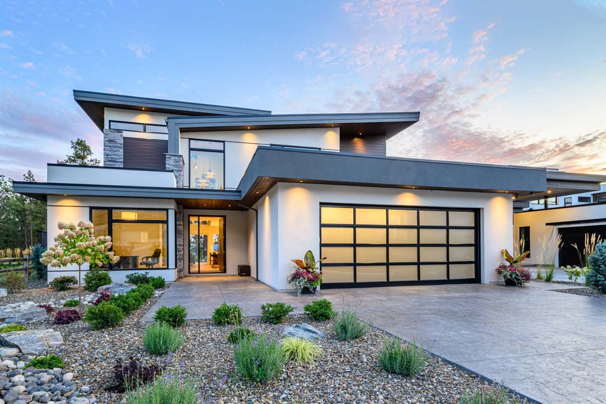 House plan exterior. Modern style with flat rooflines, a glass garage door, and large windows. Includes landscaping with small plants and rocks.