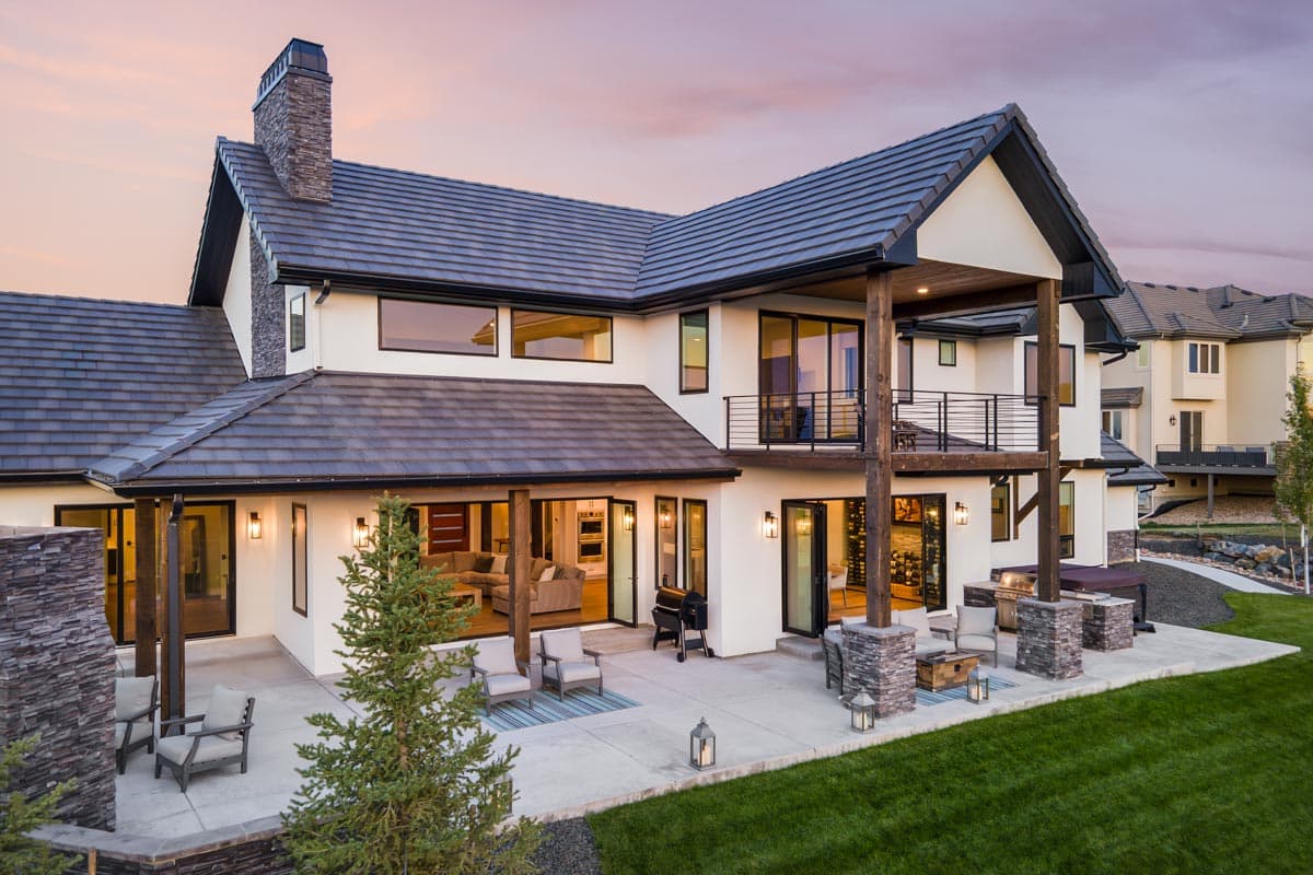 House plan exterior: two-story modern home with expansive patio, upper balcony, stone chimney, and large windows.