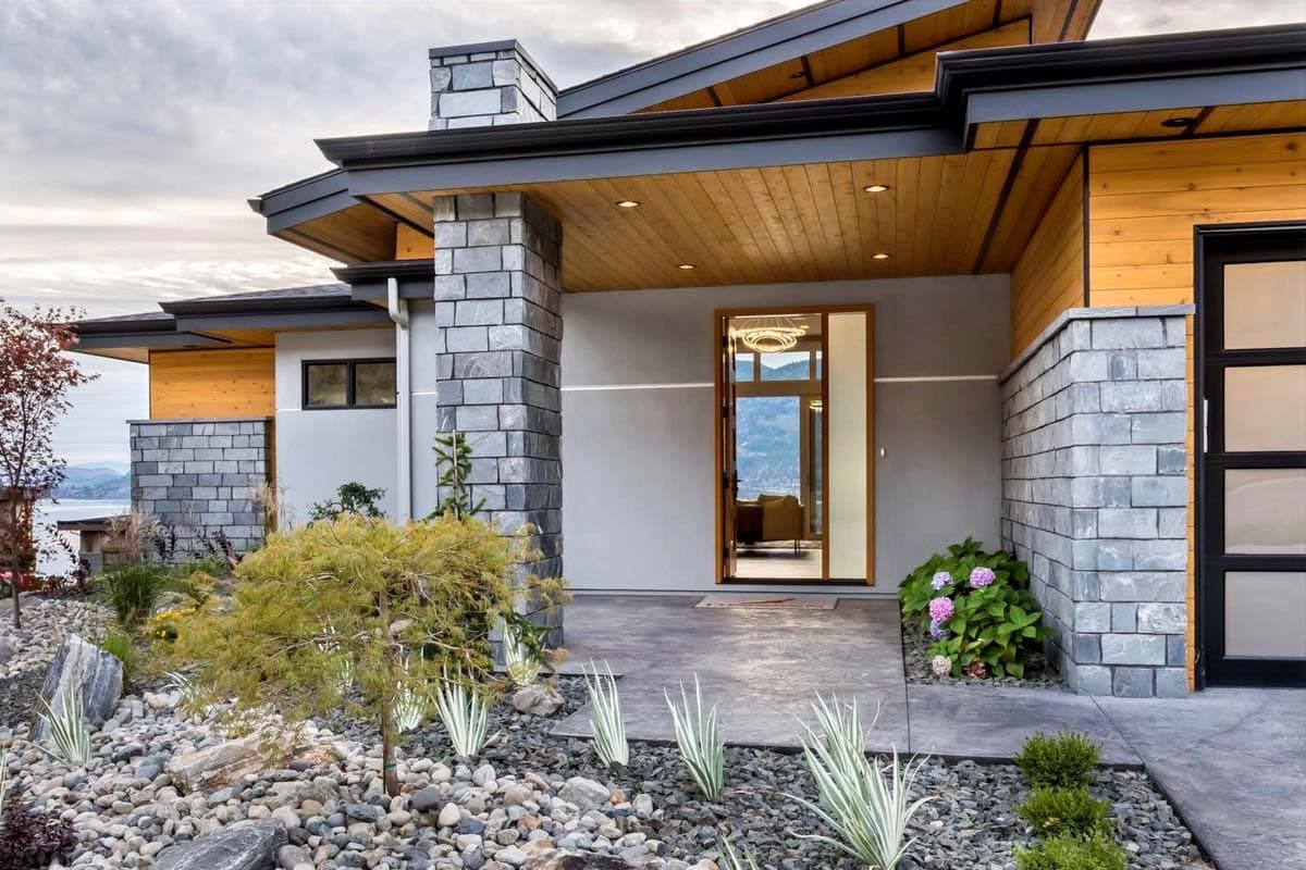 Modern house plan exterior with layered rooflines, stacked stone chimney, wood soffits, and glass entry door.