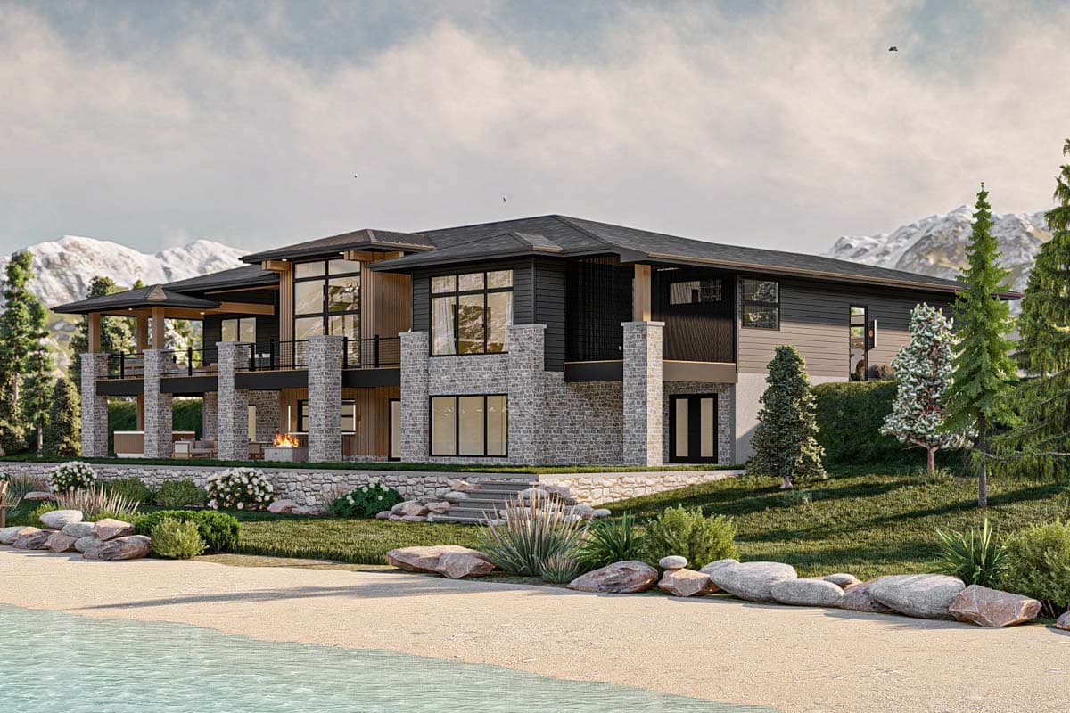 A modern, multi-level home with stone and wood accents sits beside a lake with mountains in the background. The house has large windows and balconies.
