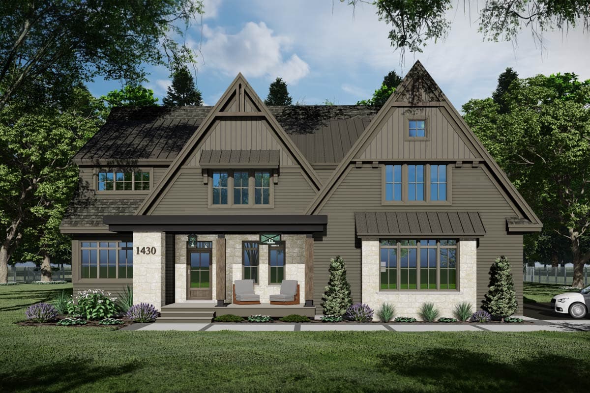 A modern, two-story house with a gabled roof, stone accents, and a covered front porch with two chairs. Lush greenery surrounds the home.