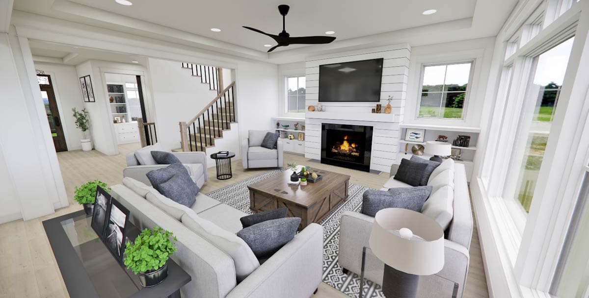 A modern living room with a fireplace, large windows, and a sectional sofa. A staircase leads upstairs, and the room is well-lit.