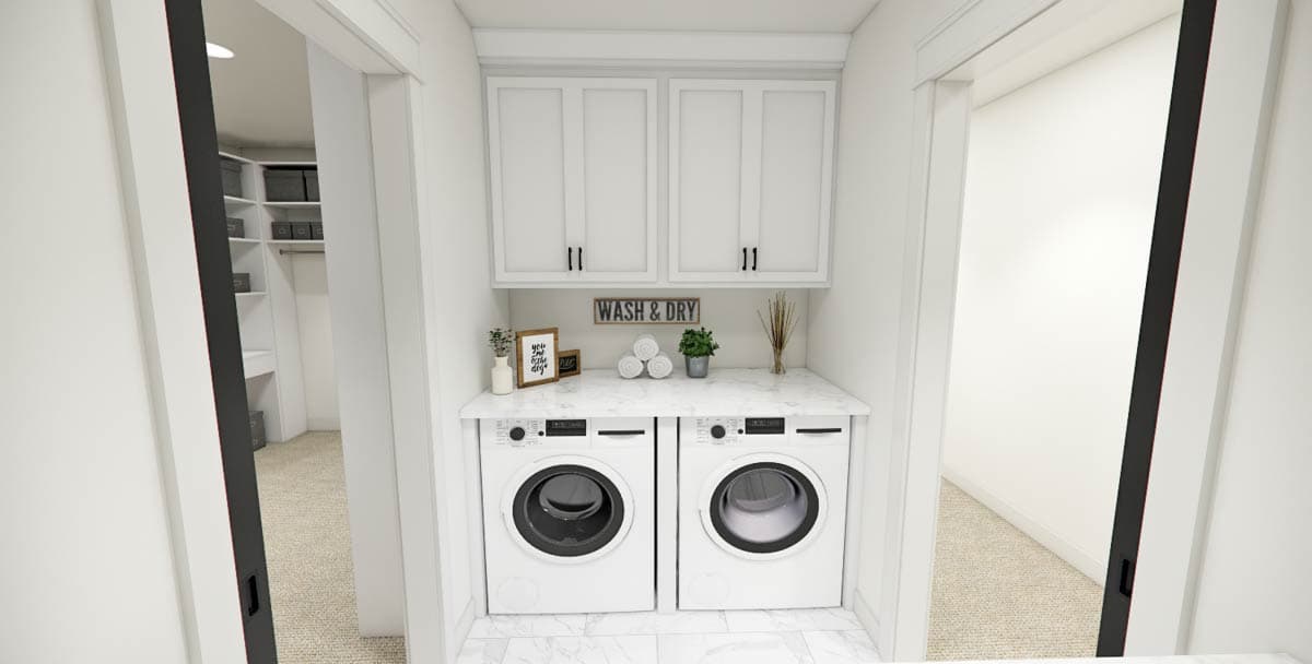 A modern laundry room with white cabinets above two front-load washing machines. Decorative elements include a "Wash & Dry" sign and a small potted plant.