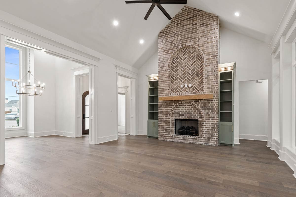 Interior view of a great room with vaulted ceiling, brick fireplace with arched herringbone pattern, built-in shelving, and large windows.