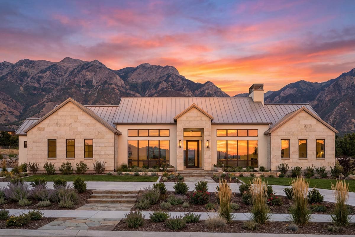 Modern house plan exterior with stone facade, metal roof, and large windows under a mountain range at sunset.