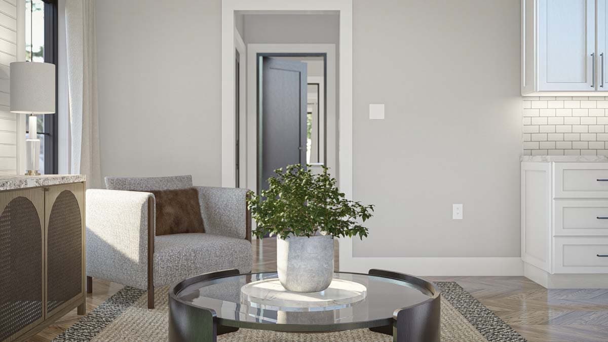 Interior view showing a living area with a gray armchair, coffee table, and doorway to another room. Kitchen cabinets visible.