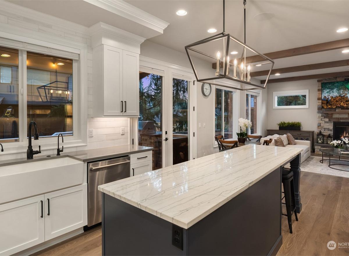 Kitchen with white cabinets, island, farmhouse sink, stainless steel appliances, and open view to living room with stone fireplace.