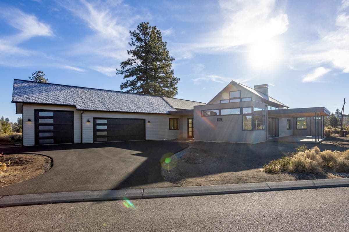 A modern, single-story house featuring a dark garage door, large windows, and a covered patio, set against a bright sky with a tall tree.