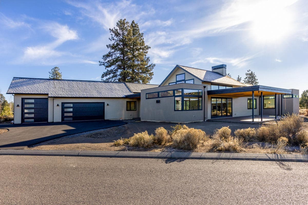 A modern home with a light exterior, black garage doors, and a covered patio area, set against a blue sky with some trees in the background.