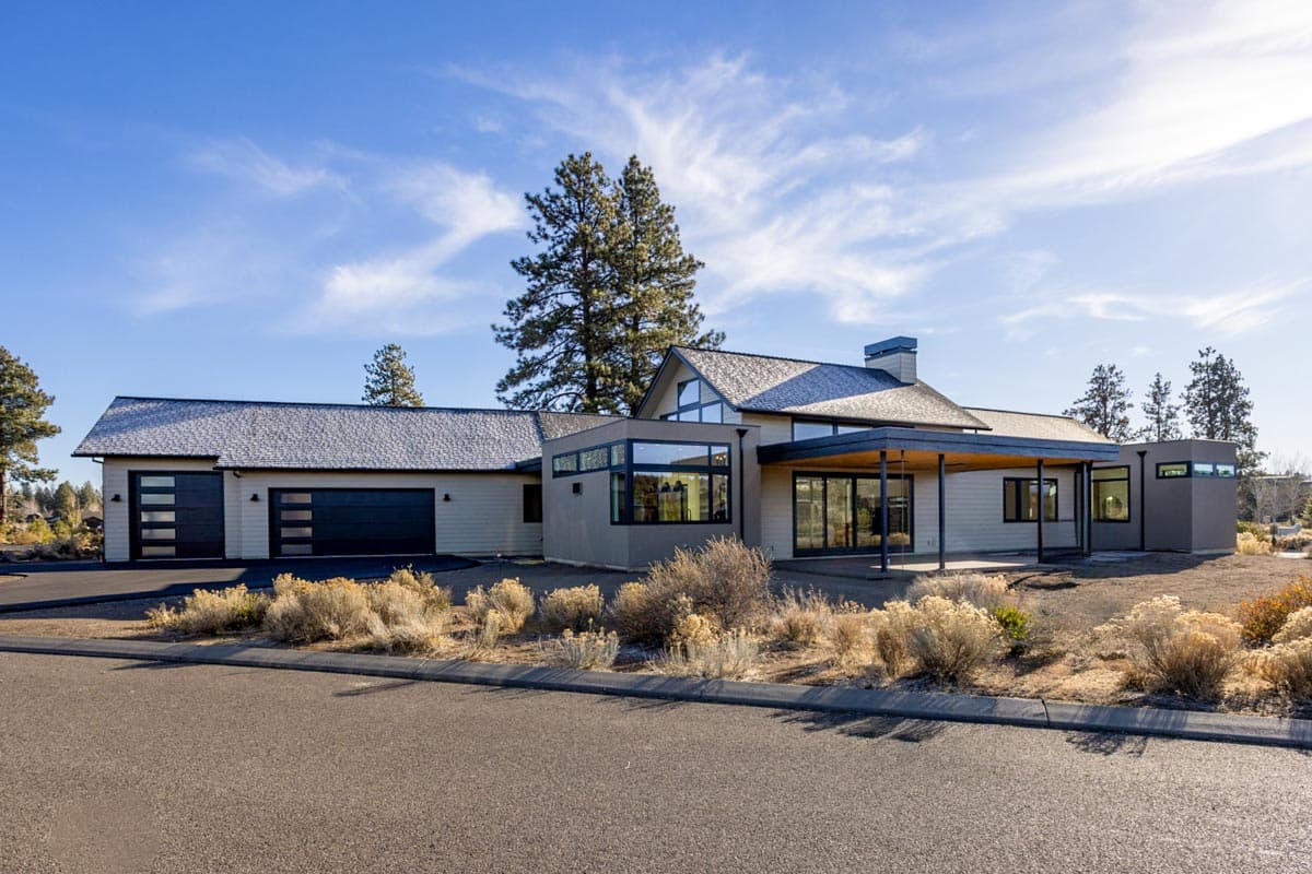A modern, beige-toned house with large windows and a covered patio under a bright blue sky. A two-car garage sits on the left.