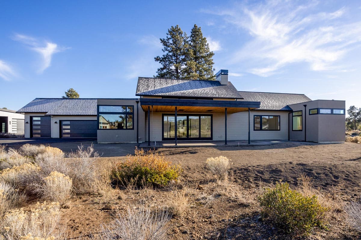 Modern, single-story house with a covered patio, large windows, and a detached garage on a sunny day. Low brush surrounds the home's foundation.