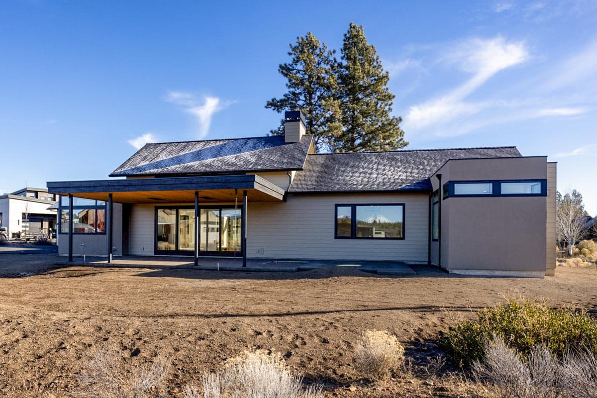 A modern, single-story home with a covered patio and large windows is set on a desert landscape, framed by a blue sky and tall trees.