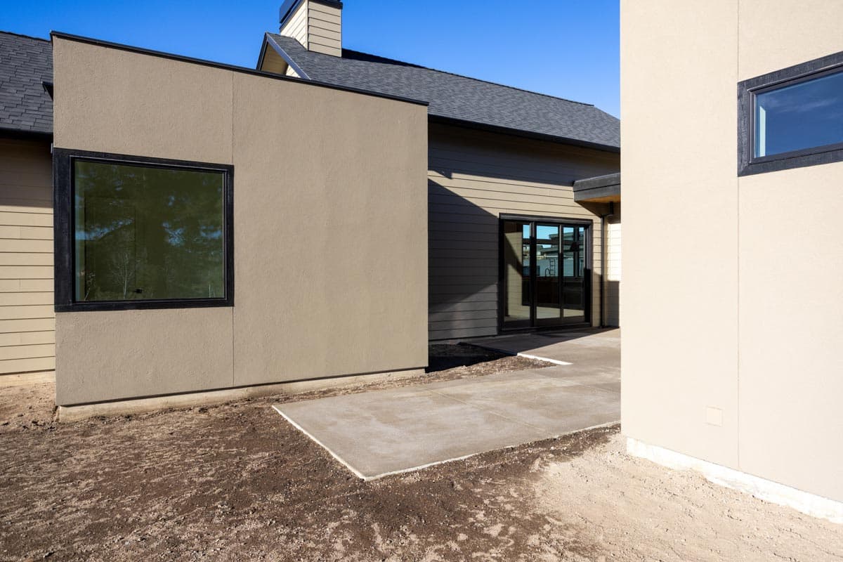 Exterior view of a modern building with beige and brown walls, large windows, and a concrete patio against a clear blue sky.