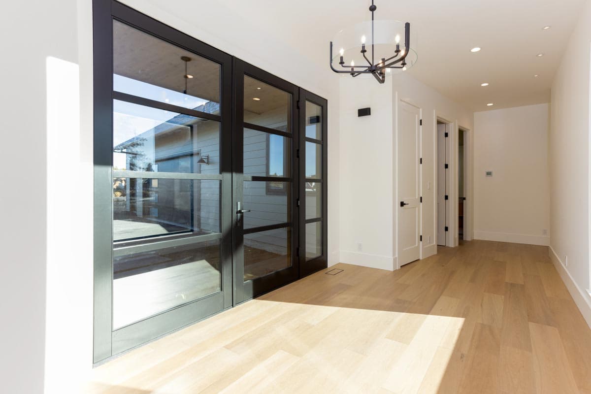 A modern home interior hallway with large, dark-framed glass doors leading outside. Natural light streams across the wood floors and walls, highlighting the space.