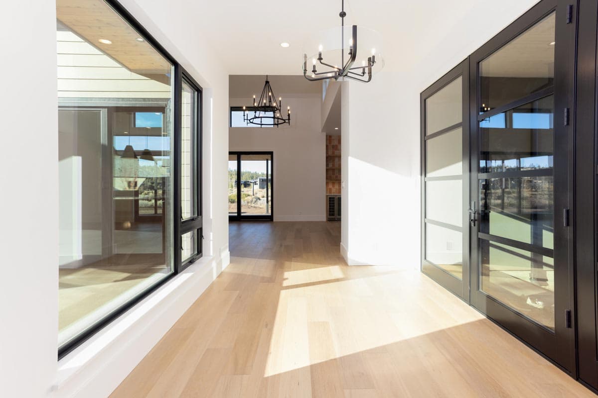 Bright, modern hallway with light wood floors, large windows reflecting the outdoors, and two black chandeliers hanging from the ceiling.