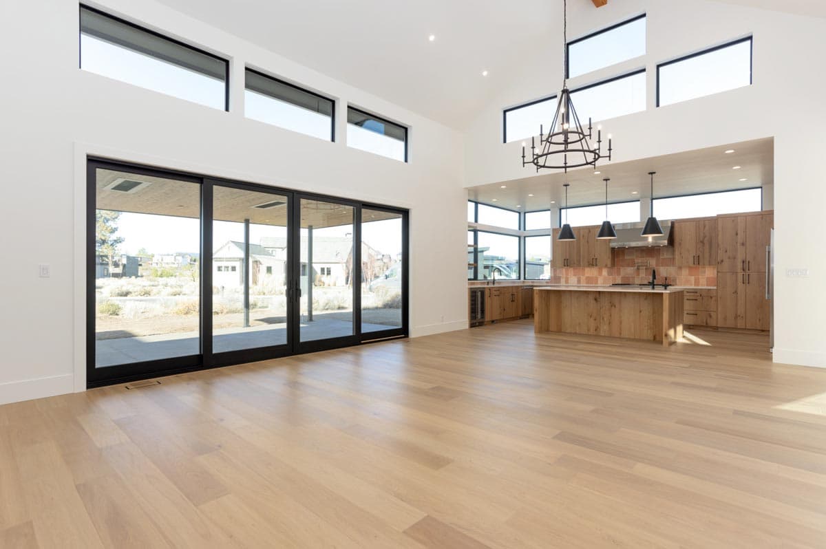 A modern, open-plan living space with large glass doors leading to a backyard. A kitchen with light wood cabinetry is visible in the distance.