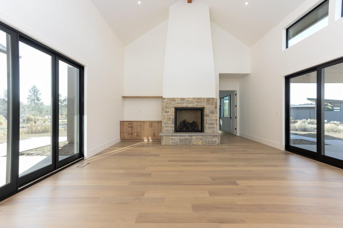 Spacious, modern living room with a stone fireplace as the focal point. Large windows provide natural light, with wooden floors and cabinets.