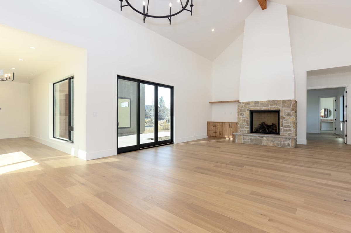 A modern, spacious living room with high ceilings, large windows, a stone fireplace, and light-colored wooden floors.