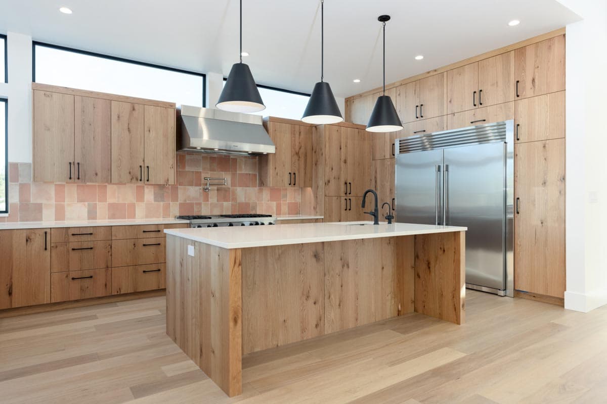 Modern kitchen with wood cabinetry and island. Overhead pendant lights hang above the island with a white countertop. Stainless steel appliances.