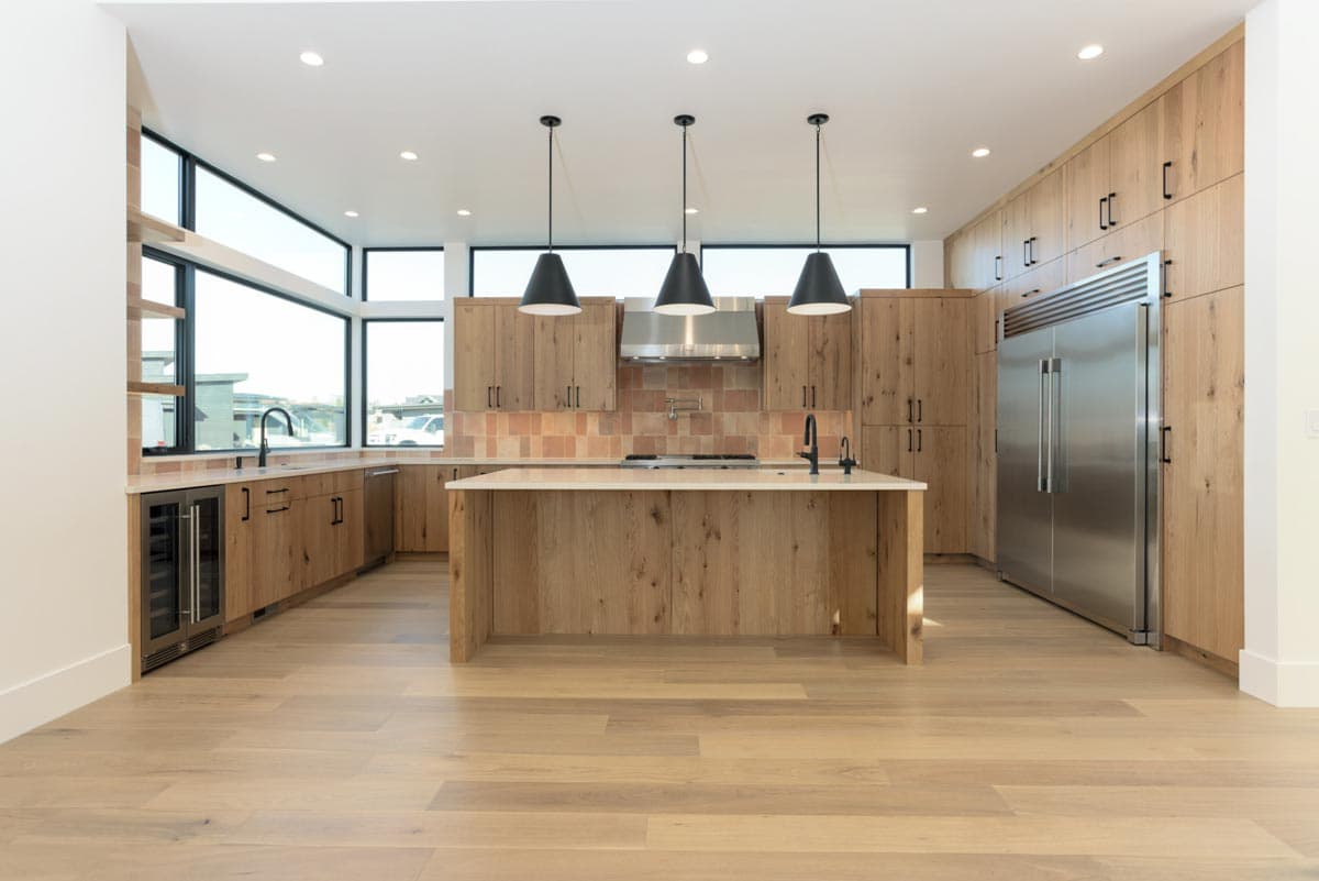 A modern kitchen with wood cabinets, island, and flooring. Stainless steel appliances contrast with the light wood, and three pendant lights hang over the island.