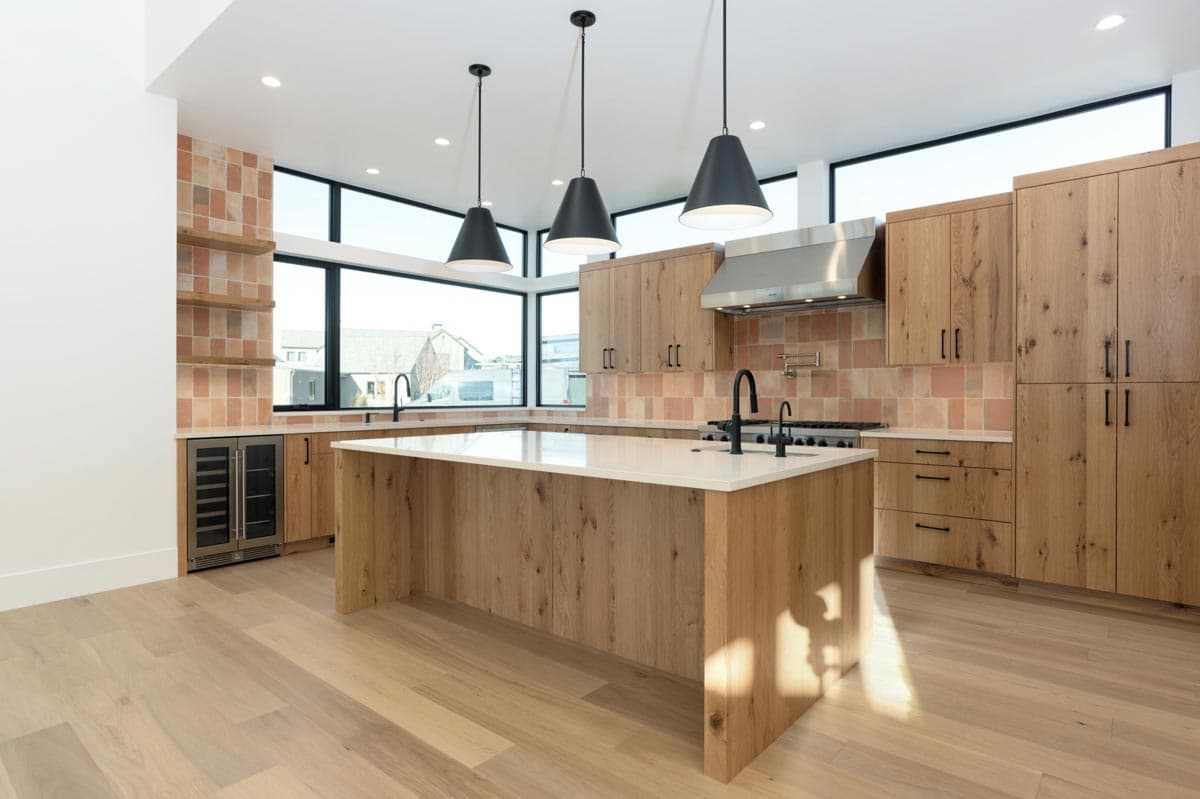 Modern kitchen with wood cabinetry, a large island, and pendant lights. A window shows the neighborhood outside, and a wine fridge is visible.