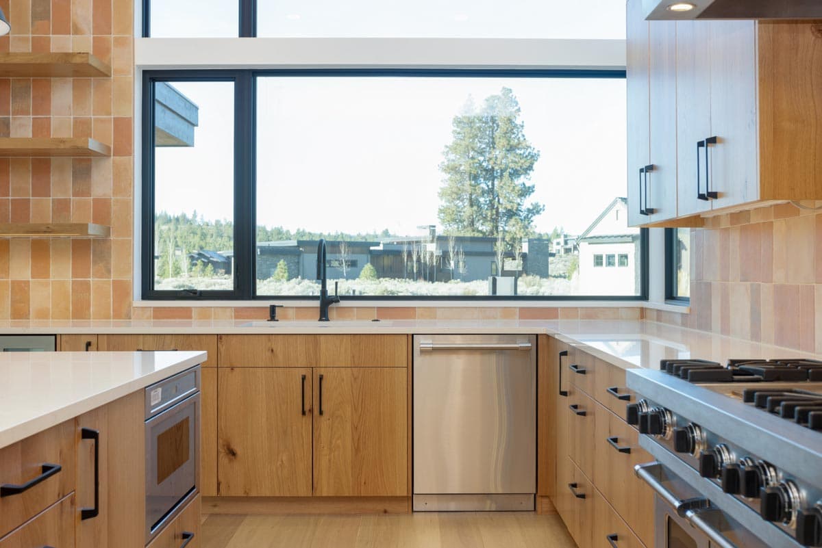 Modern kitchen with wood cabinetry, stainless steel appliances, and a large window overlooking a neighborhood with a large tree.