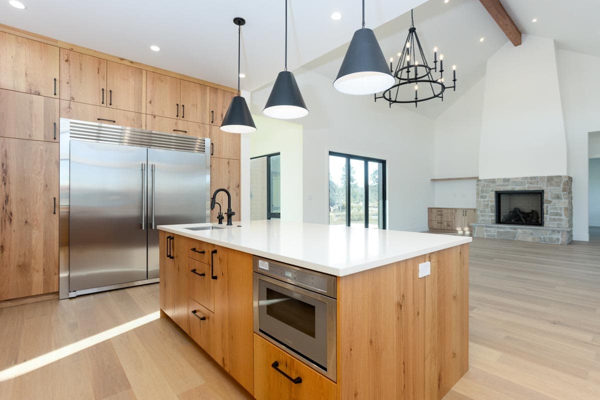 Modern kitchen with wood cabinetry, a large island with a microwave, stainless steel refrigerator, and a stone fireplace in the background.