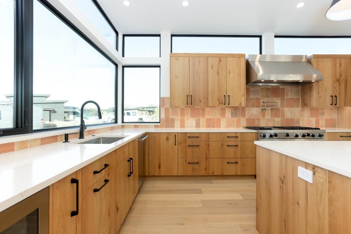 Modern kitchen with natural wood cabinets, white countertops, and large windows. Stainless steel appliances and a tiled backsplash complete the space.
