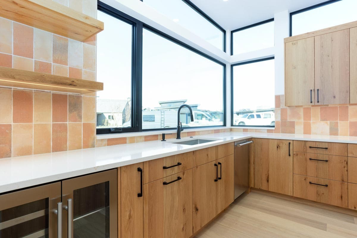 Modern kitchen with wood cabinets, white countertops, and large windows offering an expansive outdoor view. The backsplash is tiled in orange hues.