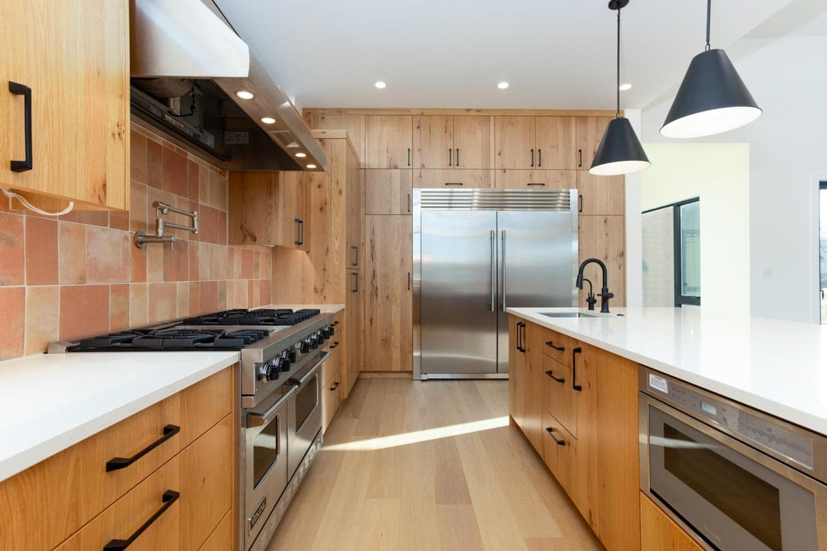 Modern kitchen with wood cabinets, stainless steel appliances, and a central island with a white countertop. Black pendant lights hang above.