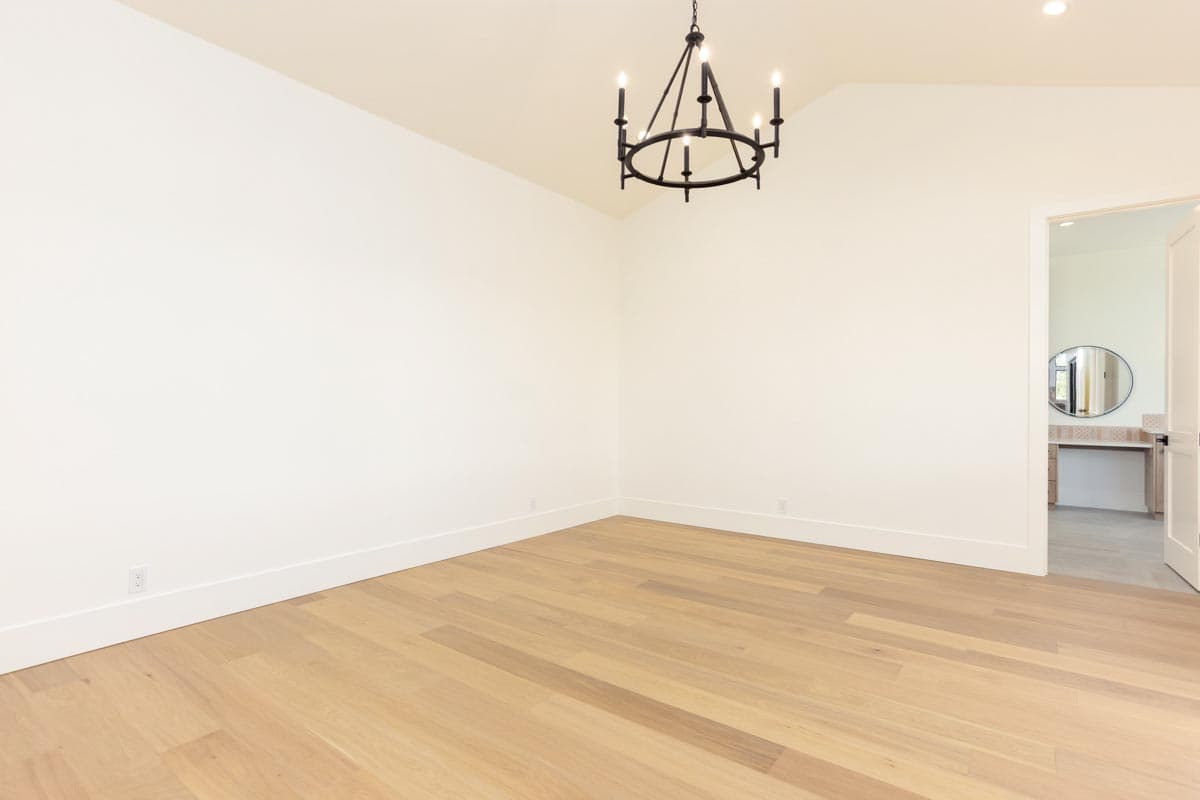 An empty room with light wood floors and white walls. A black chandelier hangs from the ceiling. A doorway reveals a vanity with a mirror.