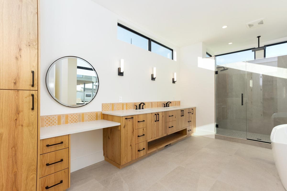 A modern, well-lit bathroom with a double vanity, a round mirror, and a glass-enclosed shower. Wooden cabinets provide storage.