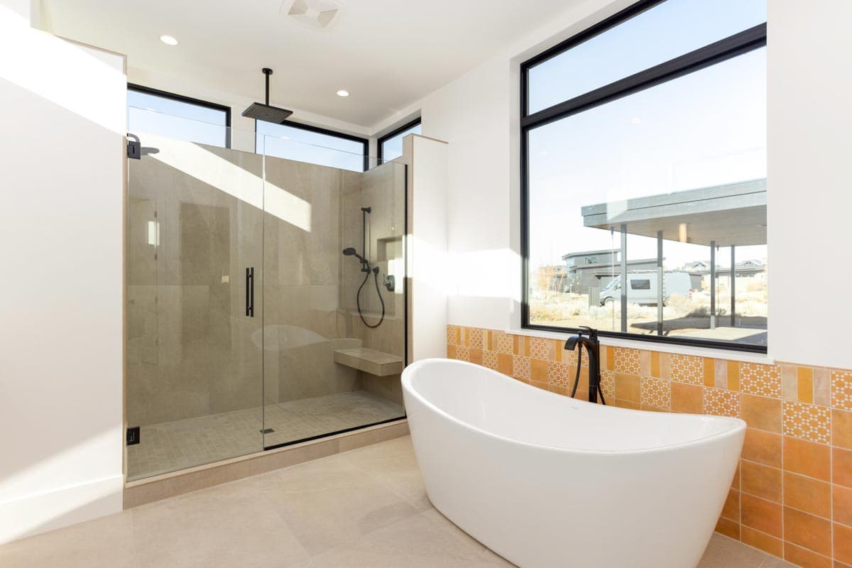 A modern bathroom with a large walk-in shower, freestanding tub, and a large window overlooking a suburban landscape.