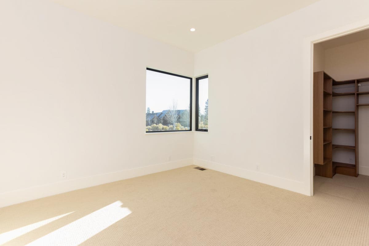 A modern, empty room with white walls, carpet flooring, and a large corner window overlooking a landscape with trees and other homes.