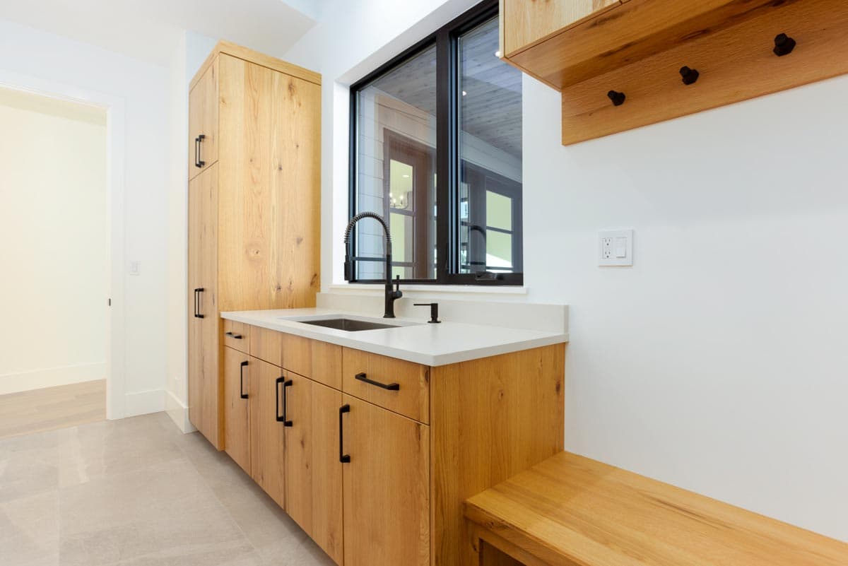 A modern laundry room with wood cabinets, white countertops, and a black faucet. A large window overlooks an outdoor space.