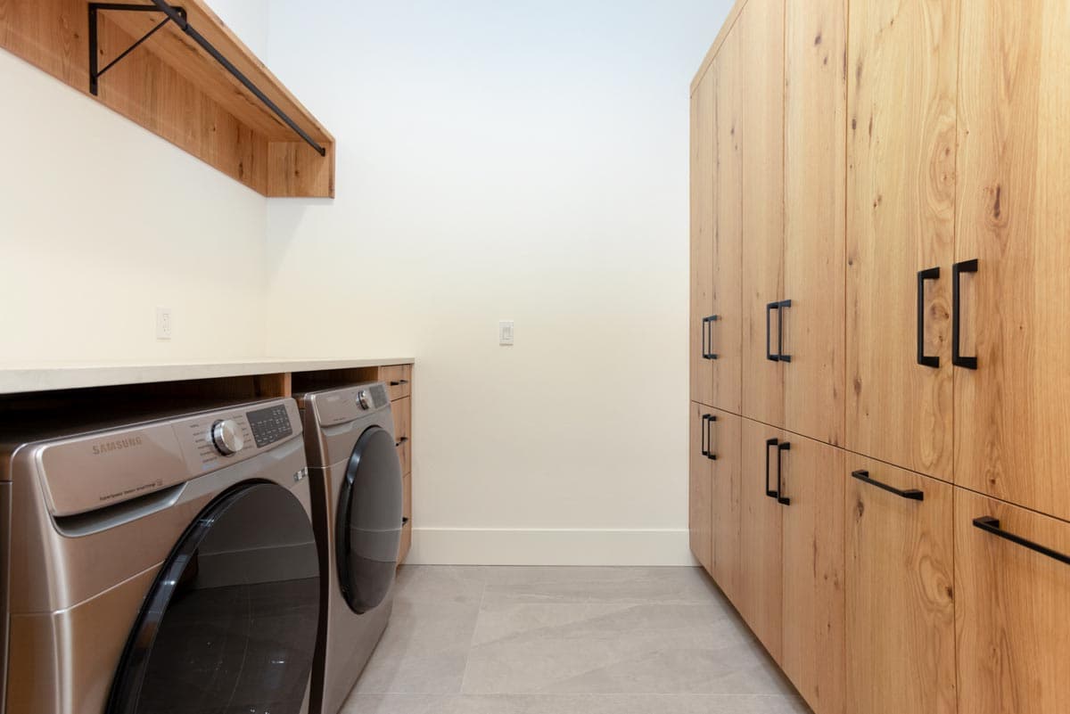 A modern laundry room with wooden cabinetry, a stainless steel washer and dryer, and light gray tiled floors.